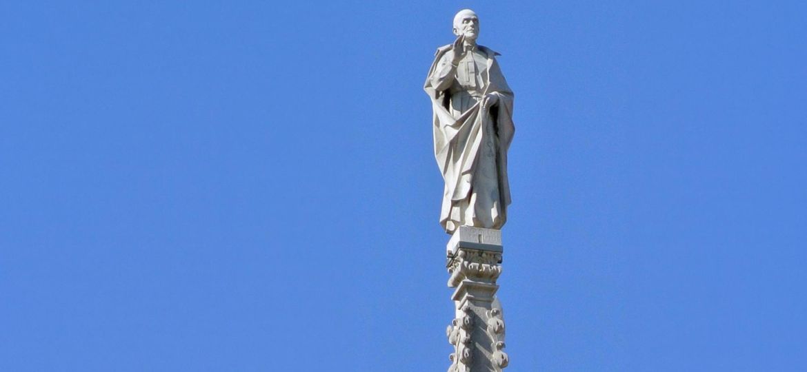 Statua bianca di San Camillo posta sul duomo di Milano. Sullo sfondo il cielo è azzurro