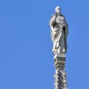 Statua bianca di San Camillo posta sul duomo di Milano. Sullo sfondo il cielo è azzurro