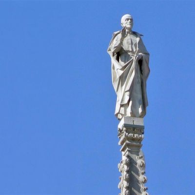 Statua bianca di San Camillo posta sul duomo di Milano. Sullo sfondo il cielo è azzurro