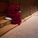Una figura vestita tutta di rosso con il cappuccio chiede l'elemosina seduta su dei gradini in strada. Non si vede la faccia.