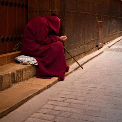 Una figura vestita tutta di rosso con il cappuccio chiede l'elemosina seduta su dei gradini in strada. Non si vede la faccia.