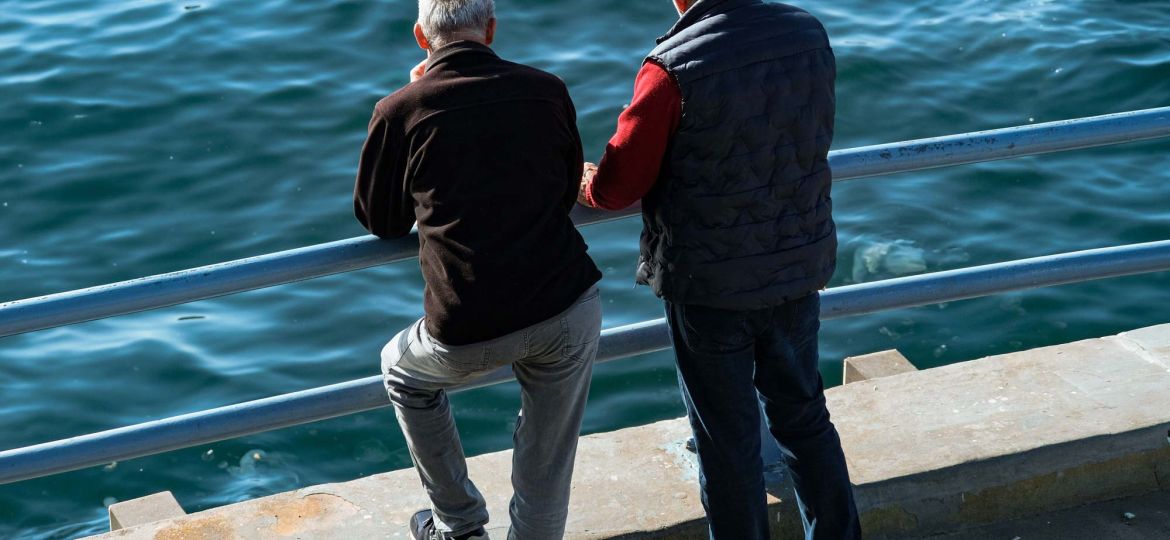 Due uomini anziani in piedi guardano il mare dalla banchina. Davanti a loro ci sono due sbarre orizzontali di protezione. Sono di spalle.
