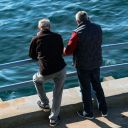 Due uomini anziani in piedi guardano il mare dalla banchina. Davanti a loro ci sono due sbarre orizzontali di protezione. Sono di spalle.