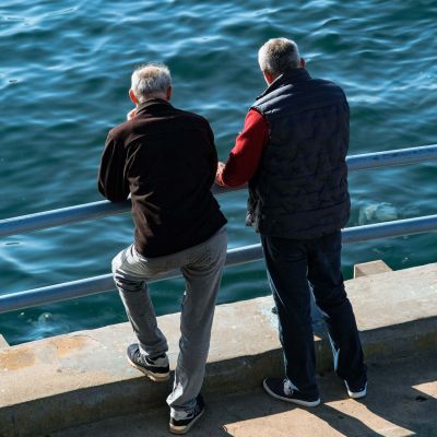 Due uomini anziani in piedi guardano il mare dalla banchina. Davanti a loro ci sono due sbarre orizzontali di protezione. Sono di spalle.